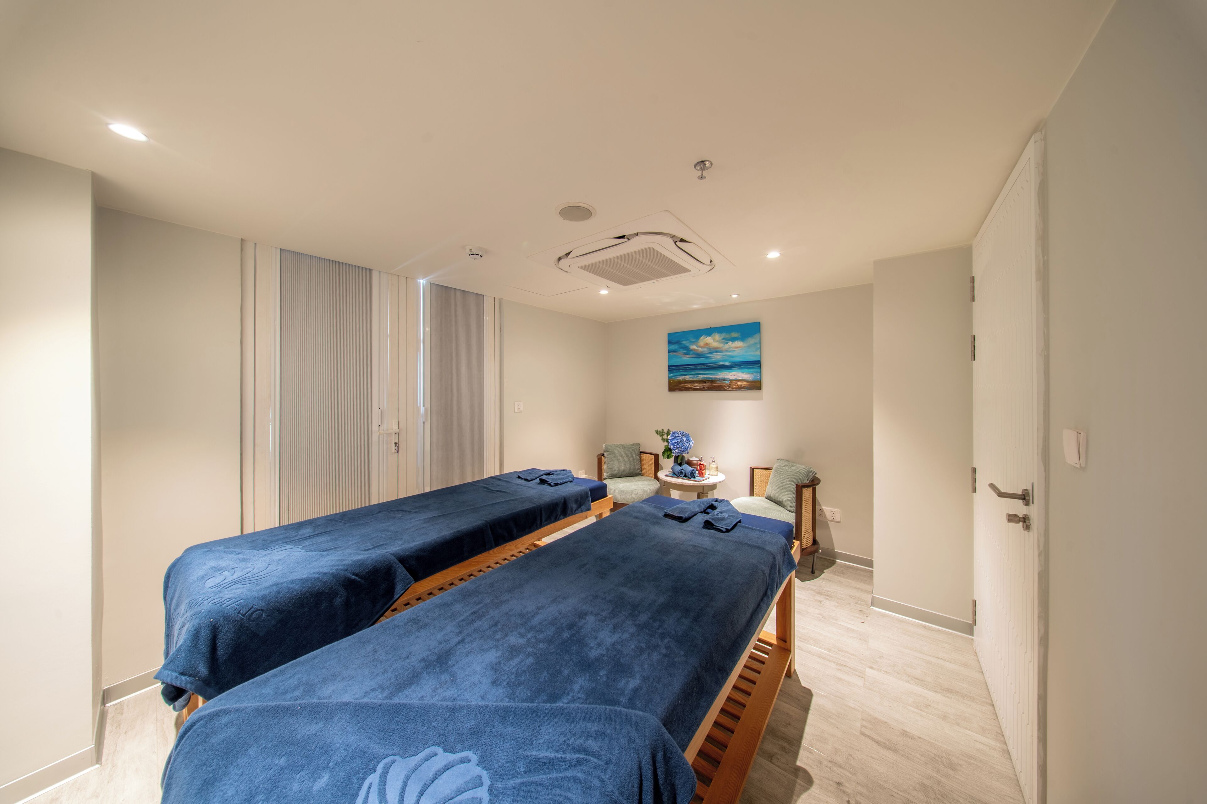 Um quarto com duas macas de massagem, uma mesa pequena com um vaso e um quadro na parede.