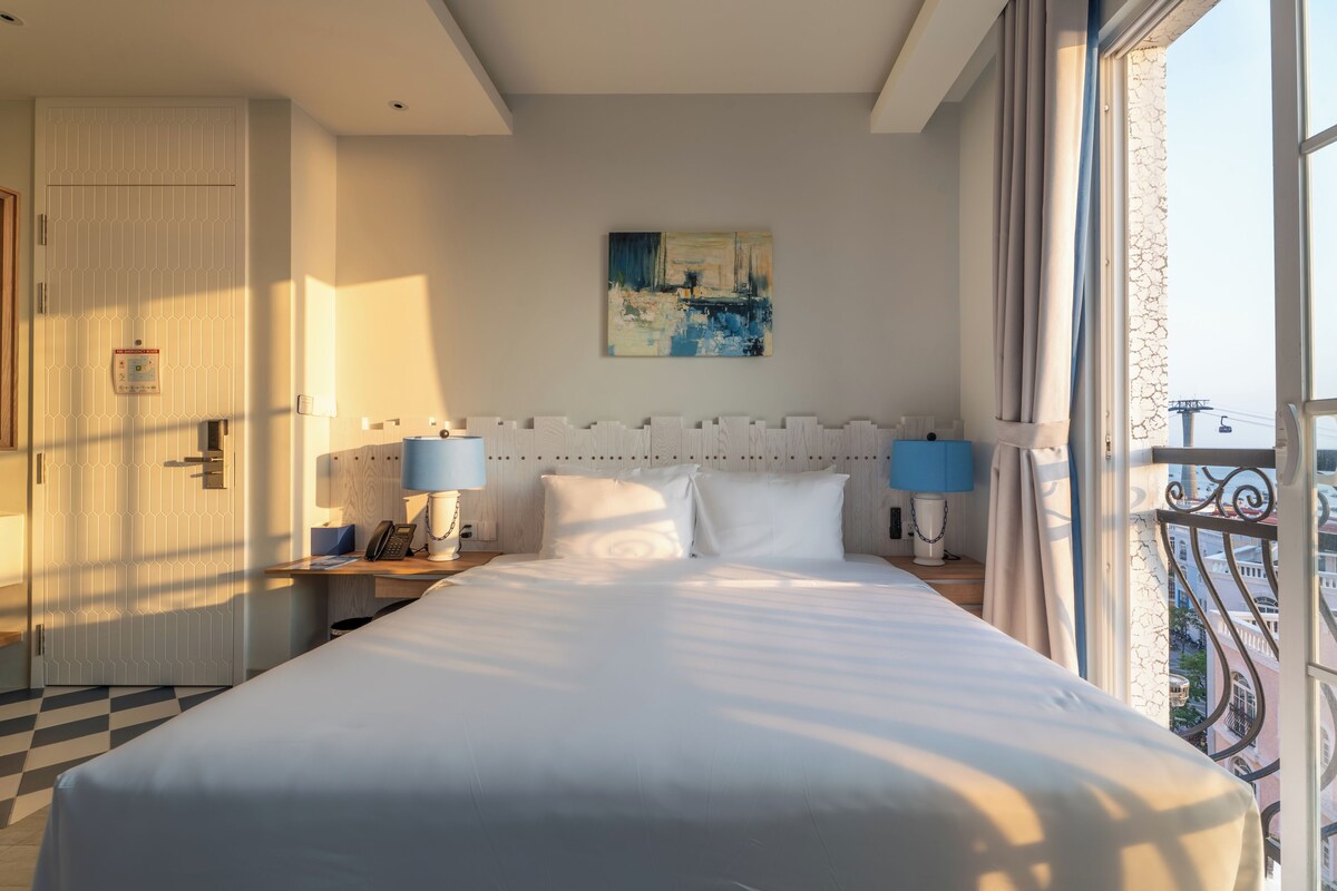 A hotel room with a bed, two bedside tables with lamps, a painting on the wall, and a balcony with a railing.