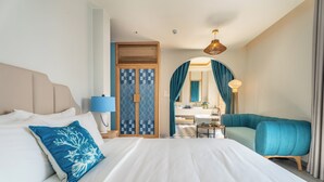 Une chambre d’hôtel avec un lit, un oreiller bleu, un canapé bleu, un bureau avec une lampe et une salle de bain visible à travers une arche.