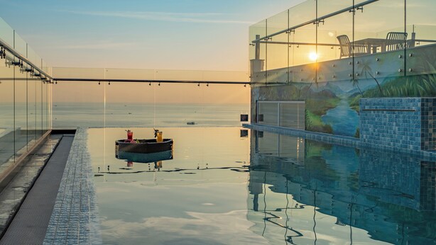 Une piscine à débordement entièrement vitrée offrant une vue sur l’océan, un escalier en verre et une fresque représentant un paysage tropical.