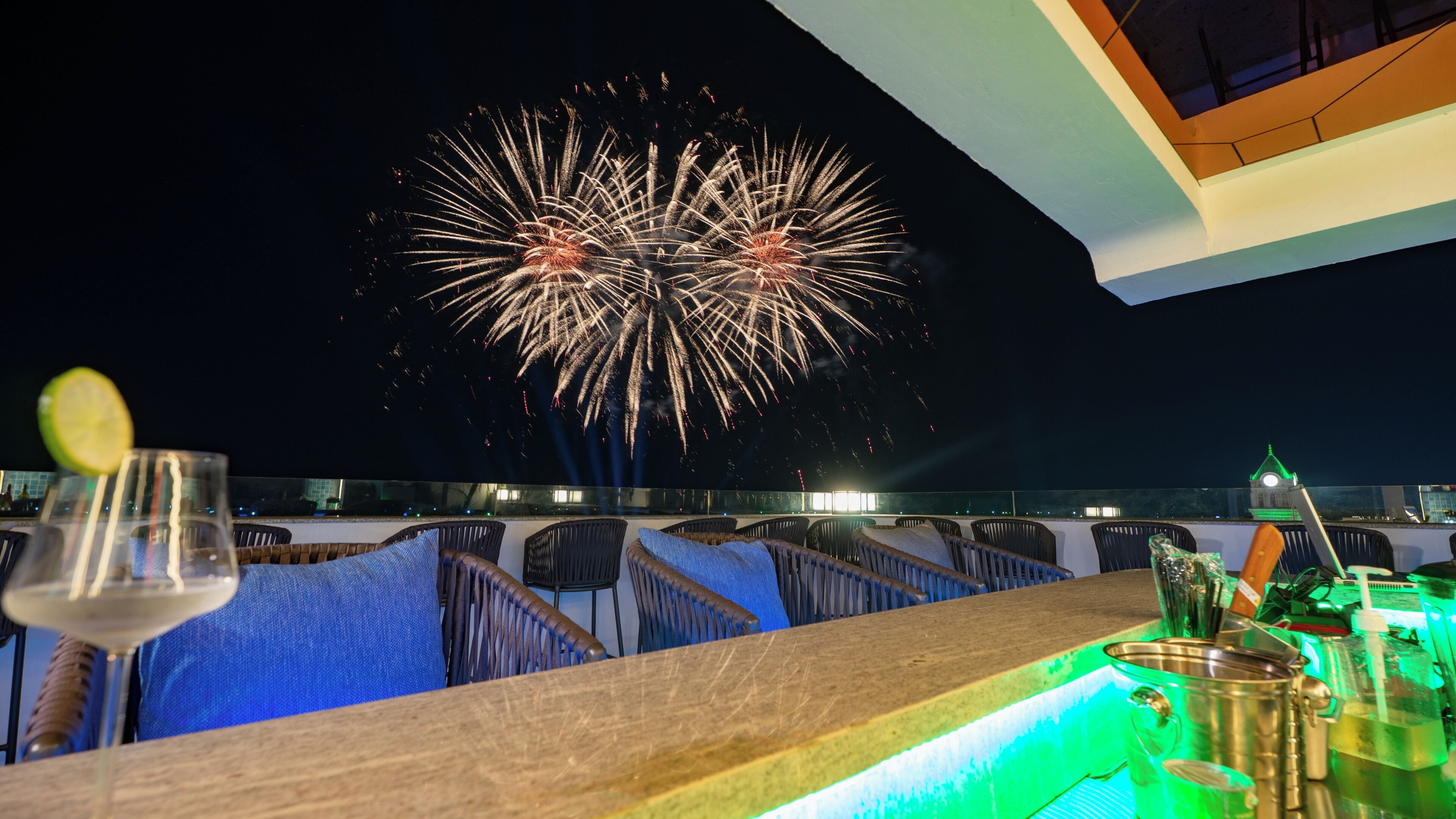 A rooftop bar with wicker chairs, a bar counter, and a view of fireworks in the night sky.