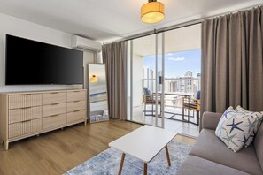A modern living room with a flat-screen TV, a sofa with a star-patterned pillow, a coffee table, and a view of the city through large windows. - Exceptionally Clean Waikiki Studio Near Beach - Ocean & Mountain Views (Honolulu)