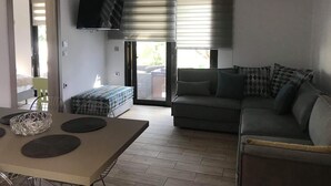 A living room with a sofa, a dining table with placemats, a TV mounted on the wall, and a sliding glass door leading to an outdoor area. - Chrysanthi Studios Apartments Pefkos (Rhodes)