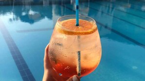 A hand holding a glass with a straw, filled with a beverage and a slice of orange, in front of a swimming pool with lounge chairs and umbrellas in the background. - Chrysanthi Studios Apartments Pefkos (Rhodes)