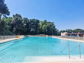 Une piscine d’un bleu éclatant, entourée d’une terrasse délimitée par une clôture, bordée d’arbres et donnant sur un bâtiment doté d’un toit en pente.