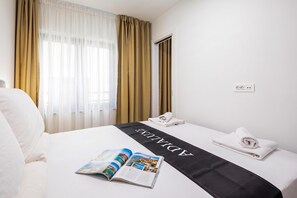 A hotel room with a bed, a window with curtains, a bedside table with a phone and towels, and a magazine on the bed. - 1 BR City Apartment Adialuxe S5, Rijeka, Croatia (Rijeka)