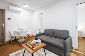 A modern living room with a grey sofa, a wooden coffee table with muffins, a white dining table with chairs, and a small side table with a lamp. - 1 BR City Apartment Adialuxe S5, Rijeka, Croatia (Rijeka)