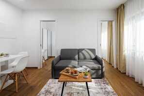 A modern living room with a grey sofa, a wooden coffee table with a magazine and pastries, a white desk with a chair, and a large window with sheer curtains. - 1 BR City Apartment Adialuxe S5, Rijeka, Croatia (Rijeka)