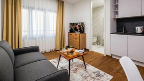 A modern living room with a sofa, a coffee table with a tray of food, a TV, and a kitchen area with cabinets and a kettle. - 1 BR City Apartment Adialuxe S5, Rijeka, Croatia (Rijeka)