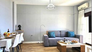 A modern living room with a grey sofa, a coffee table, and a dining area with a table set for a meal. - Albina Apartment 118 - Balcony & Sea View, Sarandë, Albania (Sarandë)
