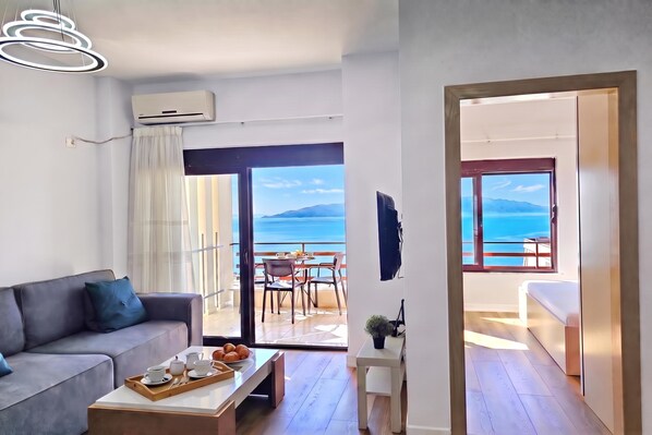 A modern living room with a sofa, a coffee table with breakfast items, and a view of the sea through large sliding doors. - Albina Apartment 118 - Balcony & Sea View, Sarandë, Albania (Sarandë)