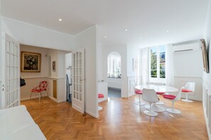 A spacious room with wooden flooring, a dining area with white round tables and chairs, and a kitchen visible through an open door. - Villa Marie by Terry's Homes (Cannes)
