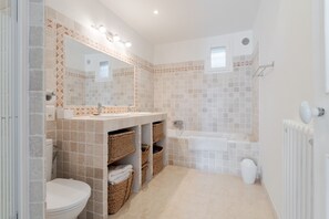 Une salle de bain avec des toilettes, un lavabo et une baignoire. On trouve des paniers en osier sous le lavabo et un porte-serviettes au-dessus de la baignoire.