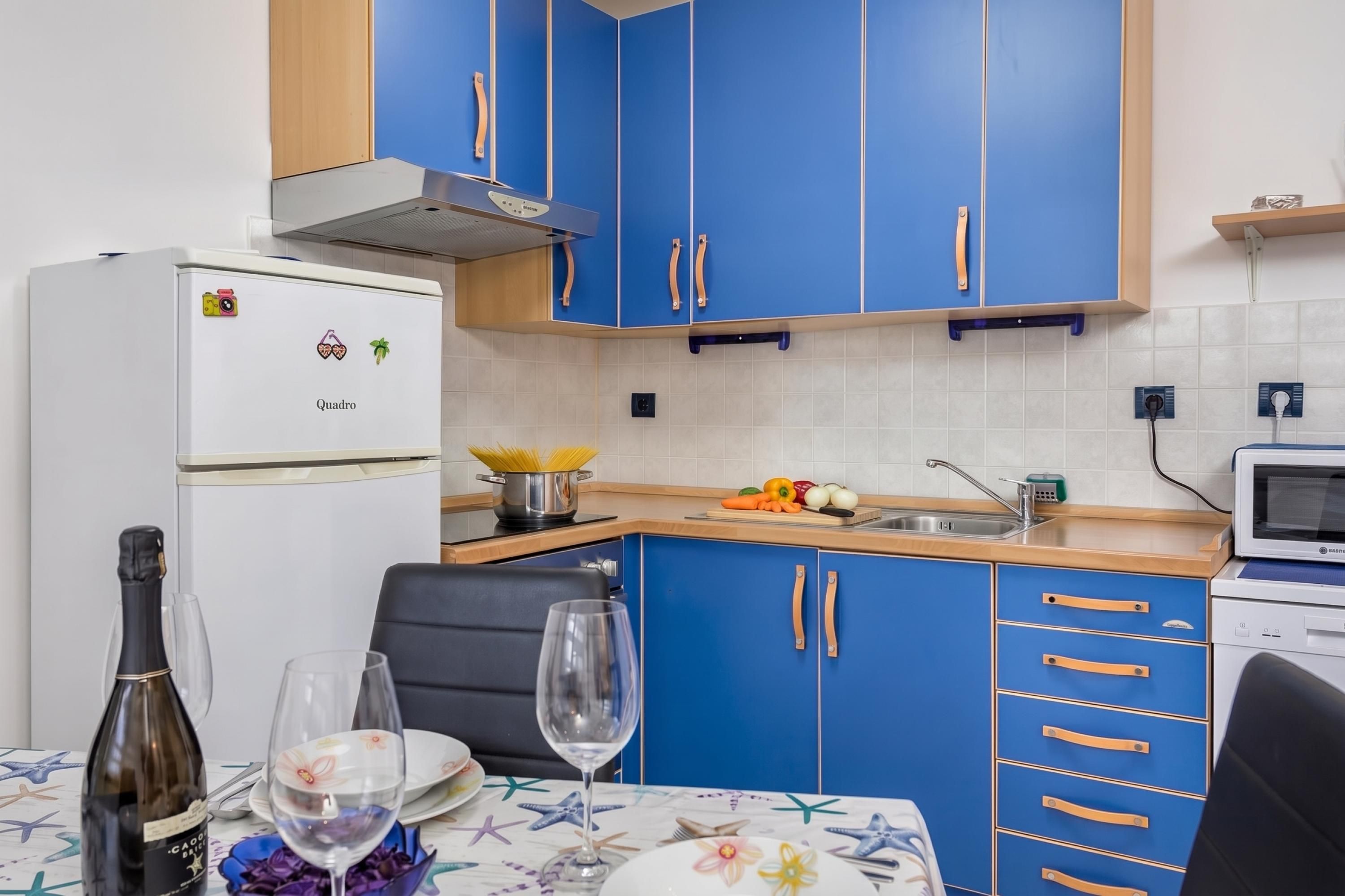 Cocina pequeña con armarios azules, un refrigerador blanco y una zona de comedor con una mesa para dos.