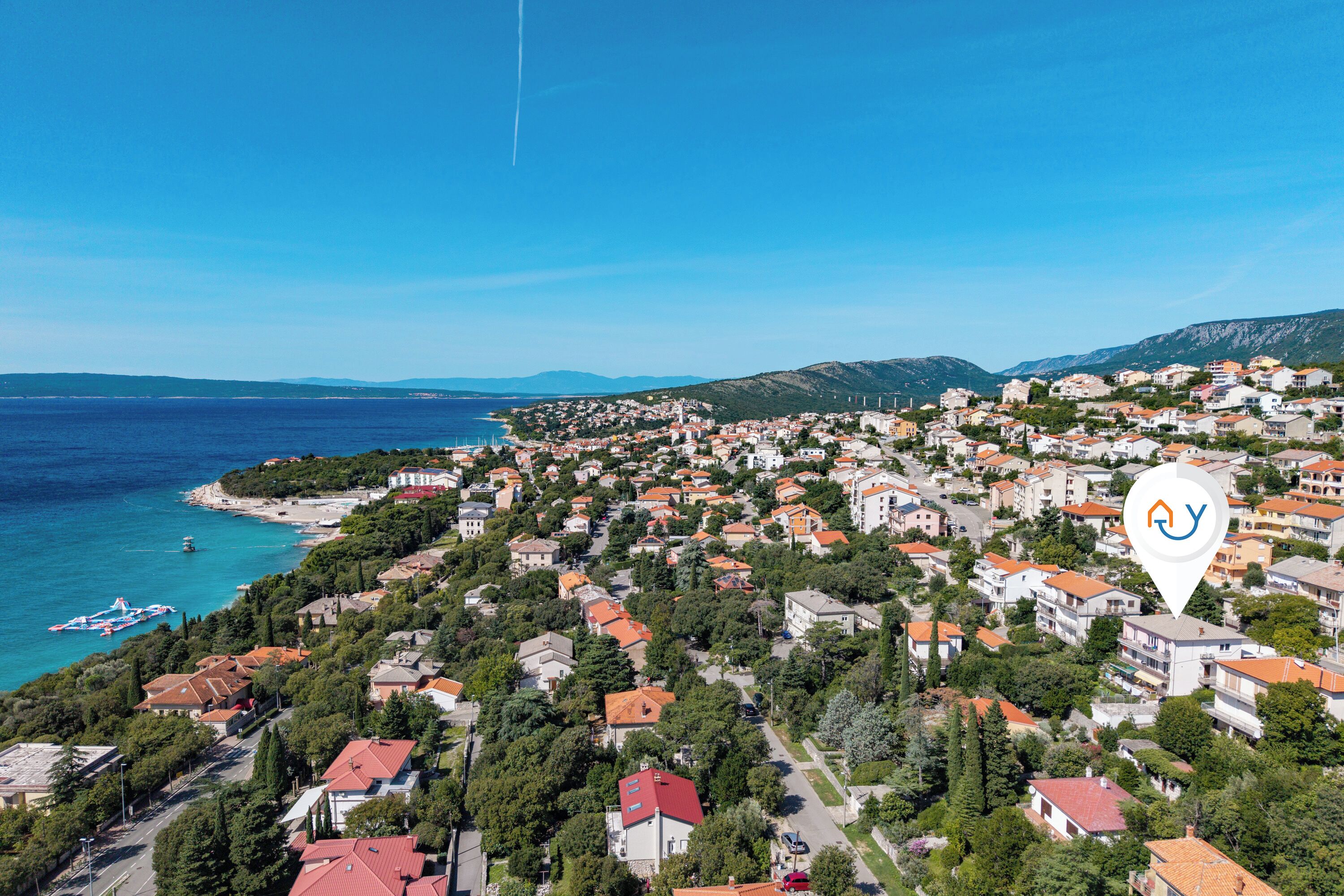 An aerial view of a coastal town with red-roofed houses, a beach, and a clear blue sky.