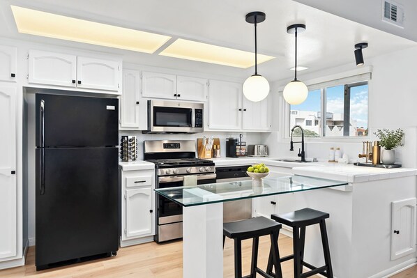 Una cocina moderna con armarios blancos, electrodomésticos de acero inoxidable, una isla de vidrio con taburetes e iluminación colgante.