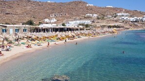 Una playa con aguas cristalinas, tumbonas y edificios blancos al fondo.