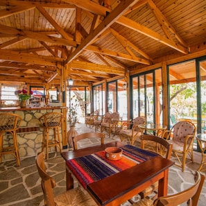 A wooden interior with a stone wall bar area, wicker furniture, and large windows. - Kastro Studios and Apartments (Ierapetra)