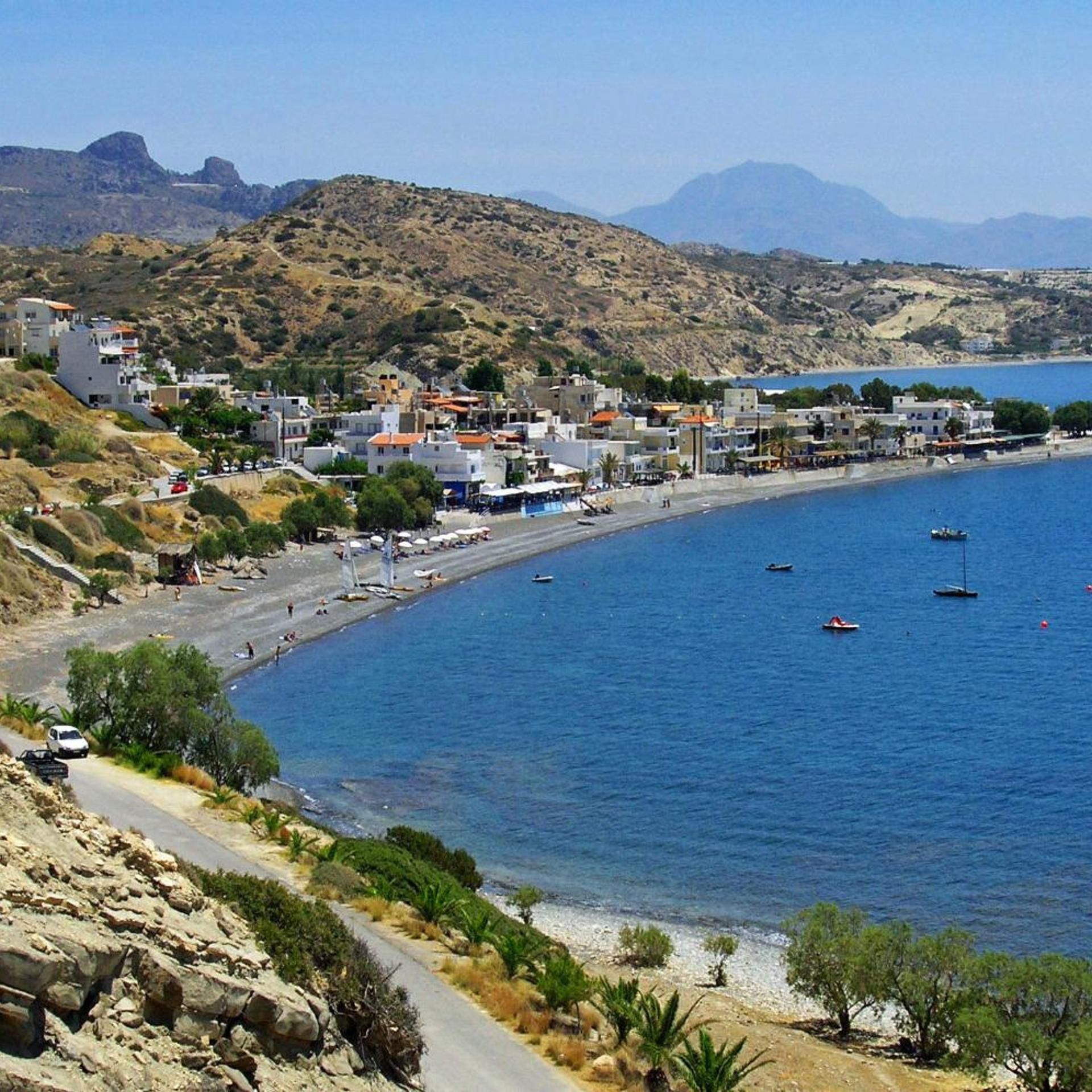 A coastal town with buildings along the waterfront, a clear blue sea, and a hilly backdrop.