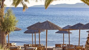 A beach with wooden lounge chairs and thatched umbrellas, overlooking a calm sea with distant mountains. - Delight Boutique Hotel & Spa (Mykonos)