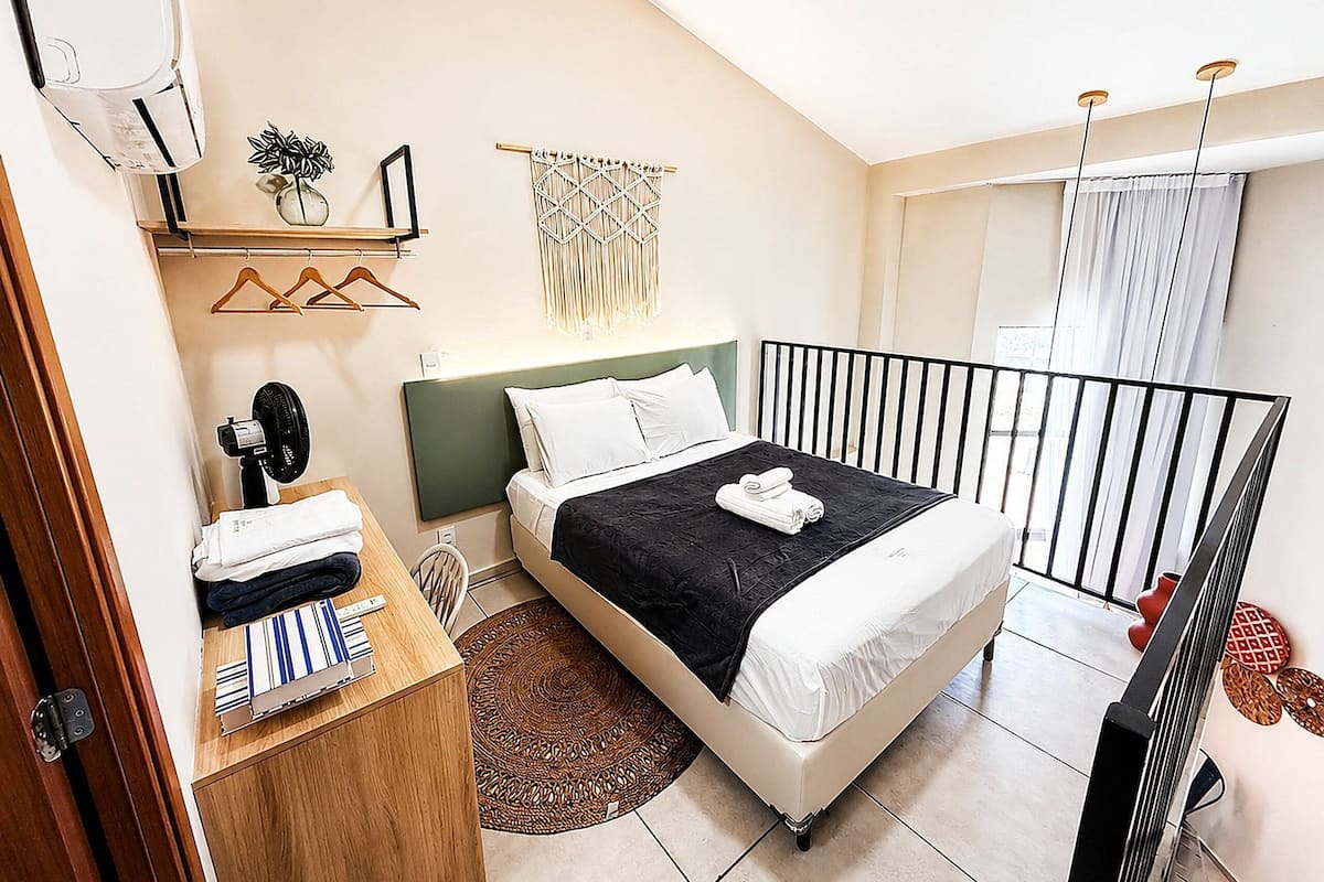 Une chambre à coucher comprenant un lit, une table de chevet, un climatiseur fixé au mur et une fenêtre avec des rideaux.