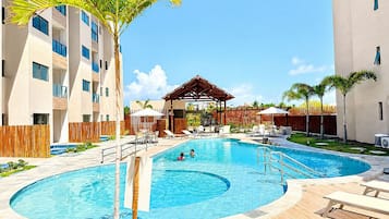 Piscina com espreguiçadeiras e guarda-sóis, cercada por edifícios com varandas e um cercado de madeira.