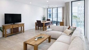Een moderne woonkamer met een grote beige hoekbank, een houten salontafel, een flatscreen televisie op een houten standaard, en een eethoek met tafel en stoelen.