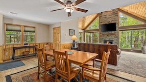 A cozy cabin interior with a dining area, a fireplace, and large windows offering a view of the forest.