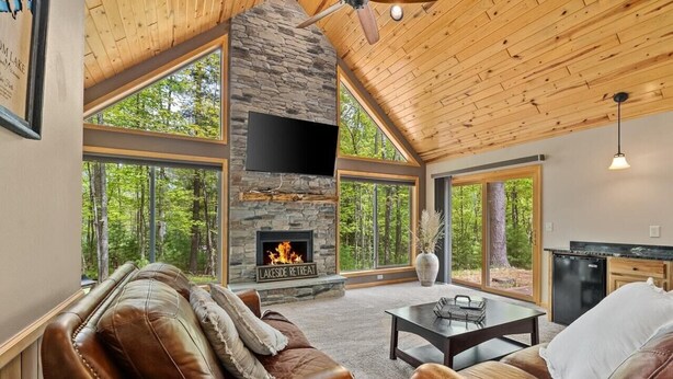 A cozy living room with a stone fireplace, leather sofas, and large windows offering a forest view.