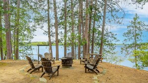 A lakeside seating area with Adirondack chairs and a fire pit, surrounded by tall pine trees.