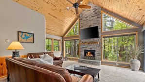 A spacious living room with a stone fireplace, leather sofa, and large windows offering a view of the forest.