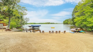 A sandy beach with several Adirondack chairs lined up facing a lake. A boat is docked on the left side of the image, and there are colorful kayaks and canoes stored on the right.