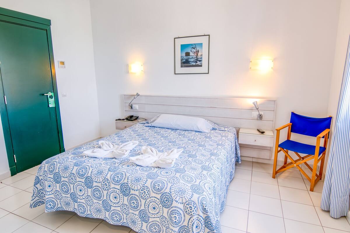 A neatly made bed with a patterned bedspread, a bedside table with a lamp, a chair, and a framed picture on the wall.