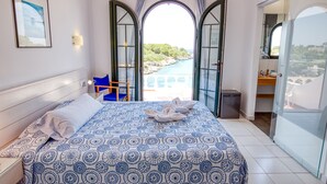 Un dormitorio con cama, silla, mesita de noche y espejo, y vistas a una piscina y vegetación a través de puertas abiertas.