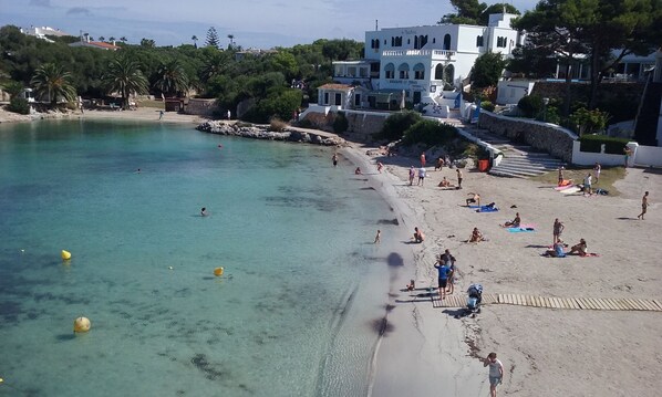 A beach with clear turquoise water, people sunbathing, and a white building with columns in the background. - Hotel Bahia (Ciutadella de Menorca)