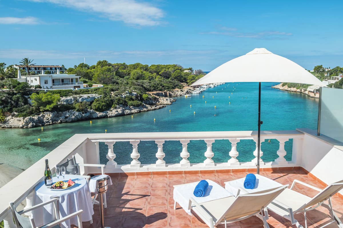 A balcony with a white umbrella, white chairs, a table with a bottle and glasses, and a view of a turquoise sea and greenery.