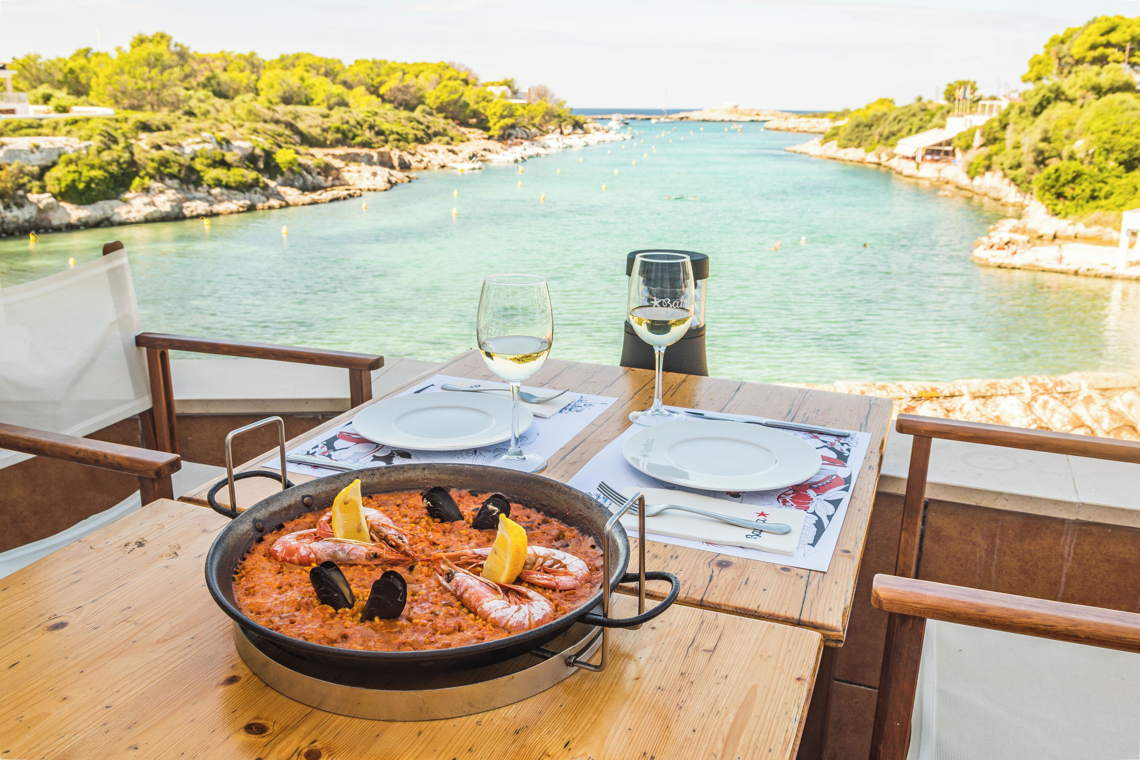 Una mesa con paella de mariscos, copas de vino y vistas a un paisaje costero.
