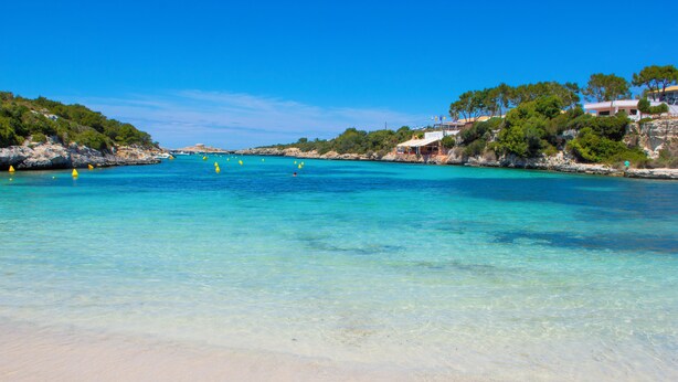 Una playa con aguas turquesas cristalinas, orilla de arena y acantilados rocosos con vegetación.