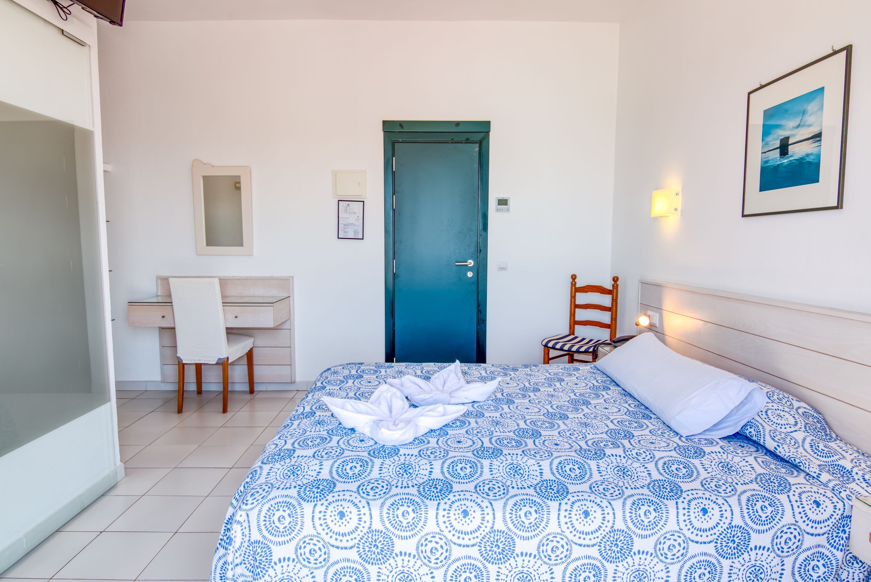 A bedroom with a bed, a desk, a chair, a door, and a wall-mounted light.