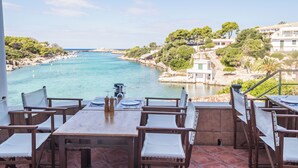 Una terraza con mesas y sillas de madera con vista a una zona costera de aguas azules cristalinas y edificios al fondo.