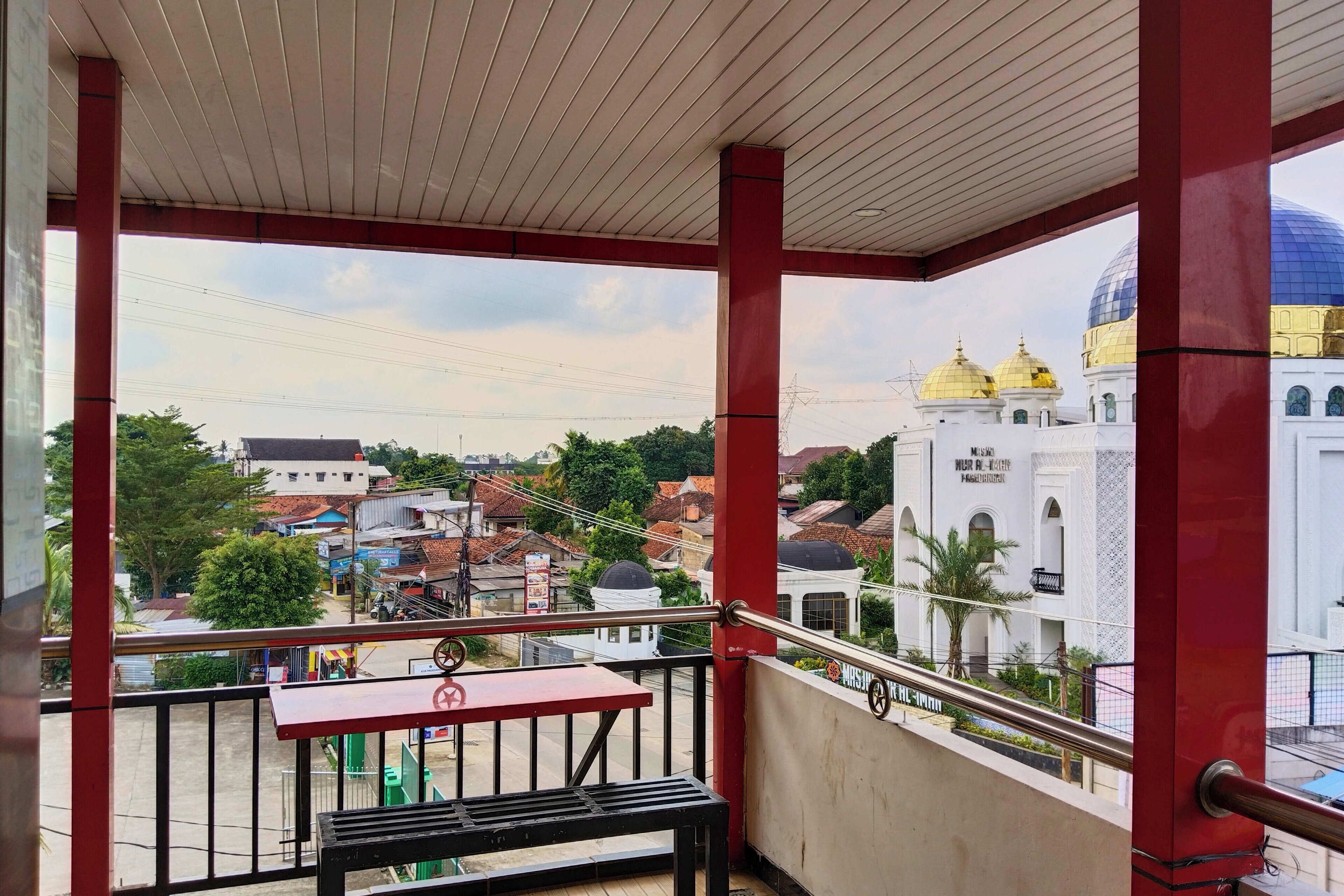A balcony with a red railing, a table, and a bench. In the background, there is a mosque with golden domes and a white building with Arabic script.