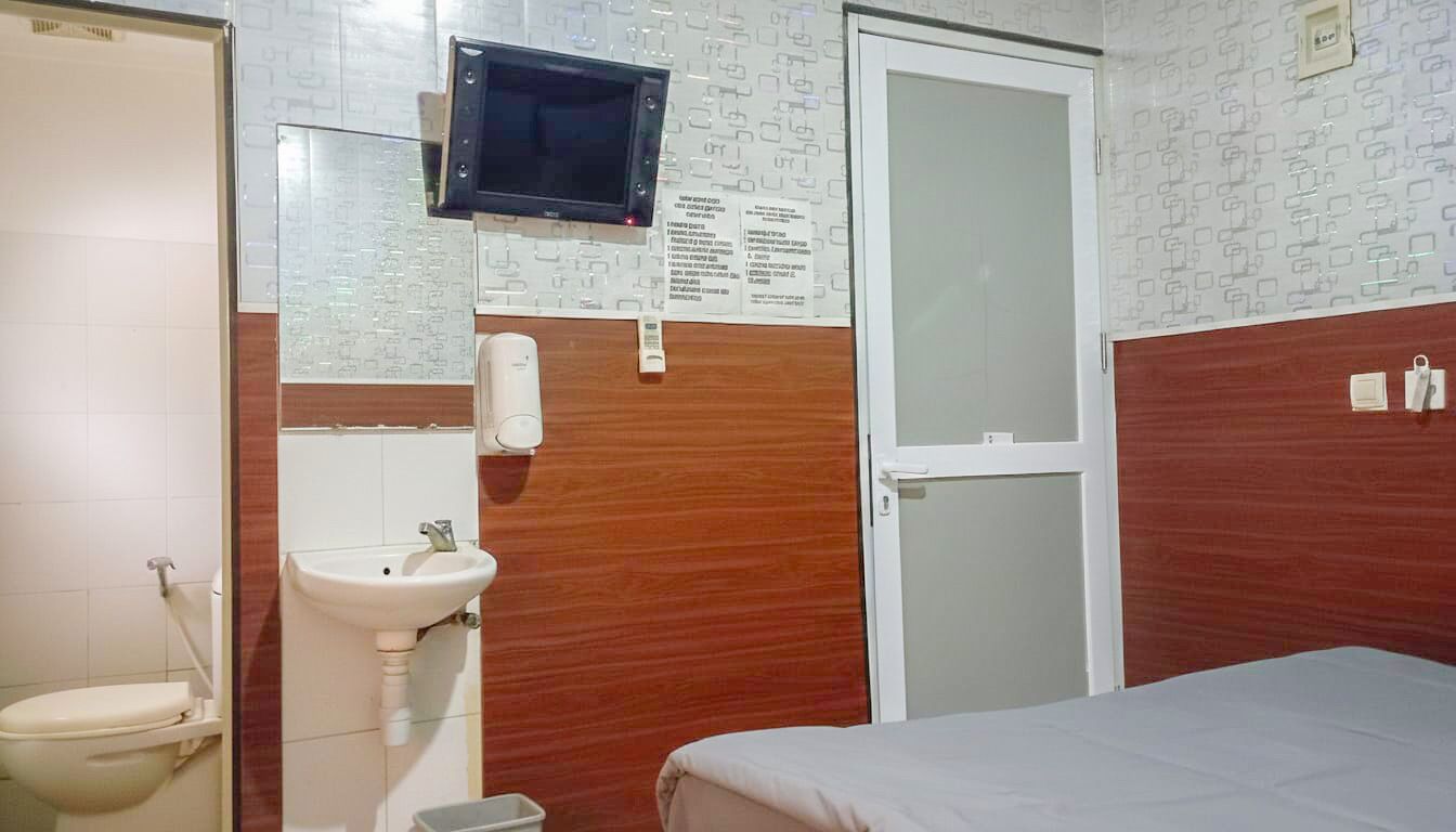 A hotel room with a wooden accent wall, a white sink, a toilet, a bed, and a television mounted on the wall.
