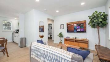 Un salon moderne avec un canapé, une table basse et un téléviseur fixé au mur. On y trouve un petit coin repas avec une table et des chaises, et une cuisine visible au fond.