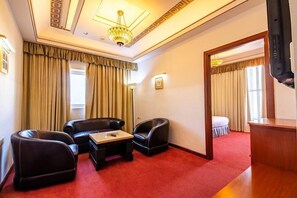 Camera d'albergo con tappeto rosso, due poltrone in pelle, un tavolino in legno, una televisione e un lampadario.