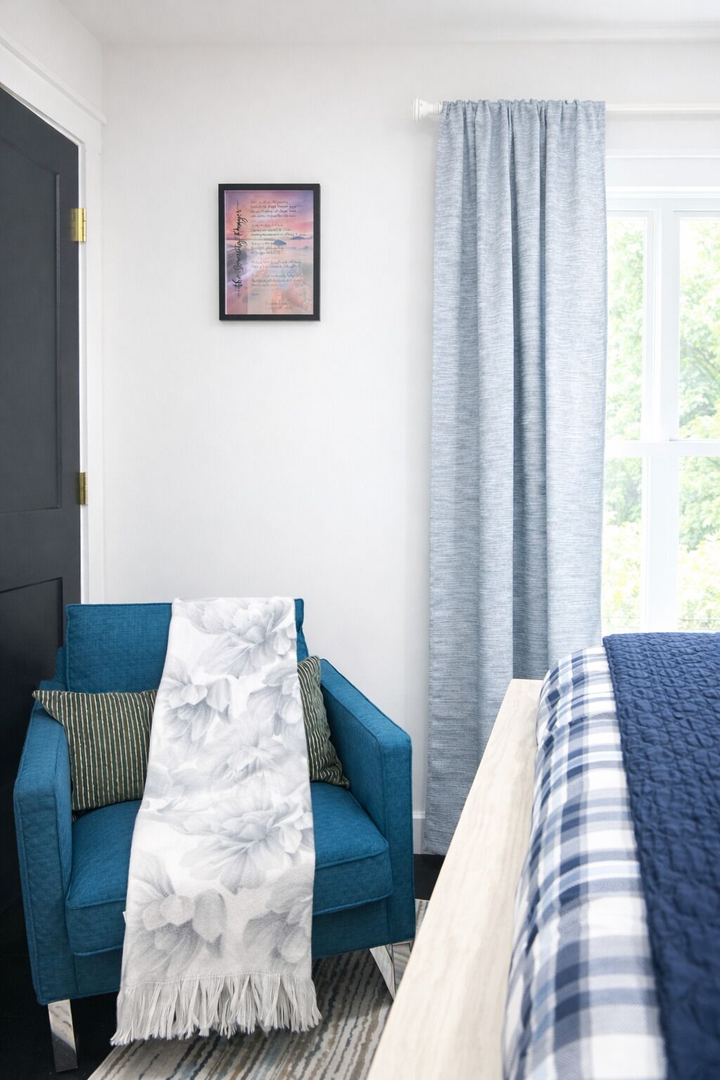 Une pièce avec un canapé bleu, un plaid blanc à motifs floraux, un tableau encadré au mur et une fenêtre avec des rideaux gris.