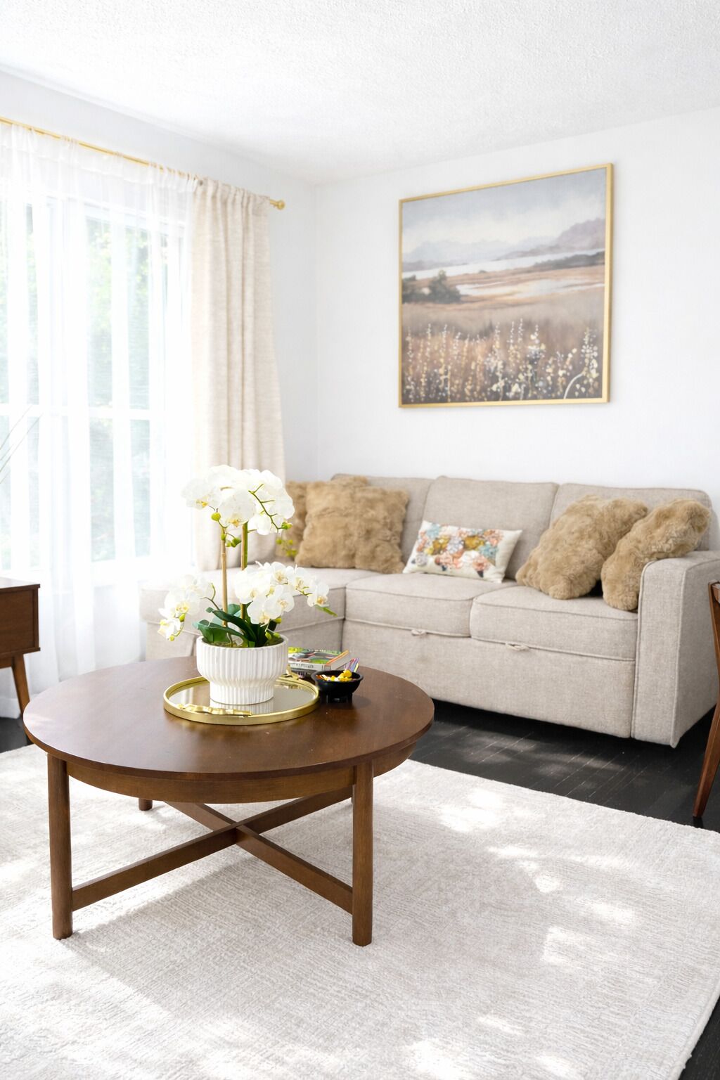 A living room with a round wooden coffee table, a beige sofa with patterned cushions, and a framed landscape painting on the wall.