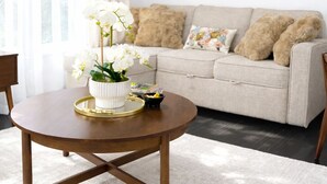 Un salon avec une table basse ronde en bois, un canapé beige agrémenté de coussins à motifs et un tableau représentant un paysage, encadré et accroché au mur.