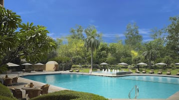 A swimming pool with lounge chairs and umbrellas, surrounded by greenery and a clear sky.