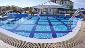 A swimming pool with a sunshade, a wooden deck, and a building in the background. - Aria Hotel (Mahala)