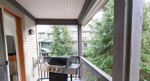 Un balcon abrité, doté d’un barbecue, d’une balustrade en bois et offrant une vue sur les maisons et les arbres des environs.